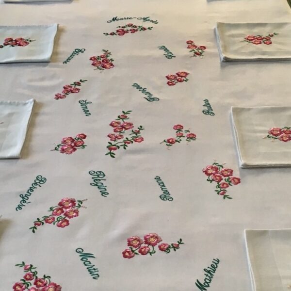 Cette nappe délicateent brodée de petites fleurs roses entrmêlées des prénoms brodés dans un camaïeu vert sur trois générations évoque les repas de famille et les dimanches partagés