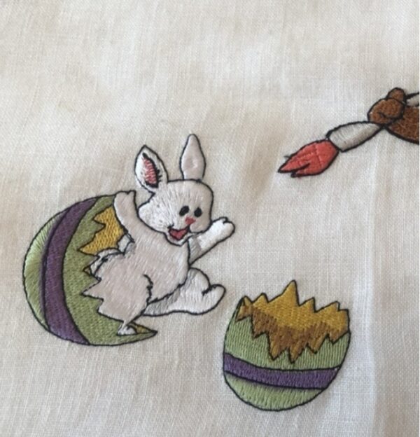 Scène de Pâques brodée représentant un grand lapin brun peignant un oeuf d'où sort un petit lapereau blanc.